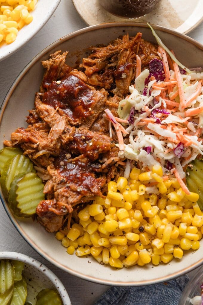 Close up of pulled BBQ pork shoulder served in a bowl with pickles, coleslaw, and corn.