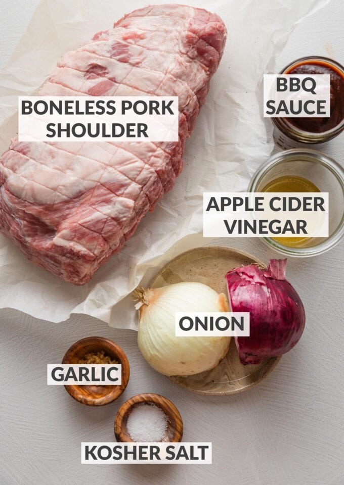 Labeled overhead photo of a boneless pork shoulder, onion, garlic, apple cider vinegar, BBQ sauce, and kosher salt.