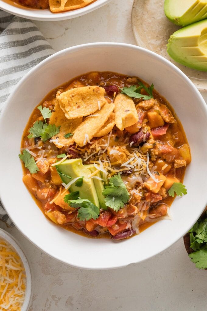 Bowl of turkey pumpkin chili topped with avocado, cilantro, shredded cheese, and corn chips.