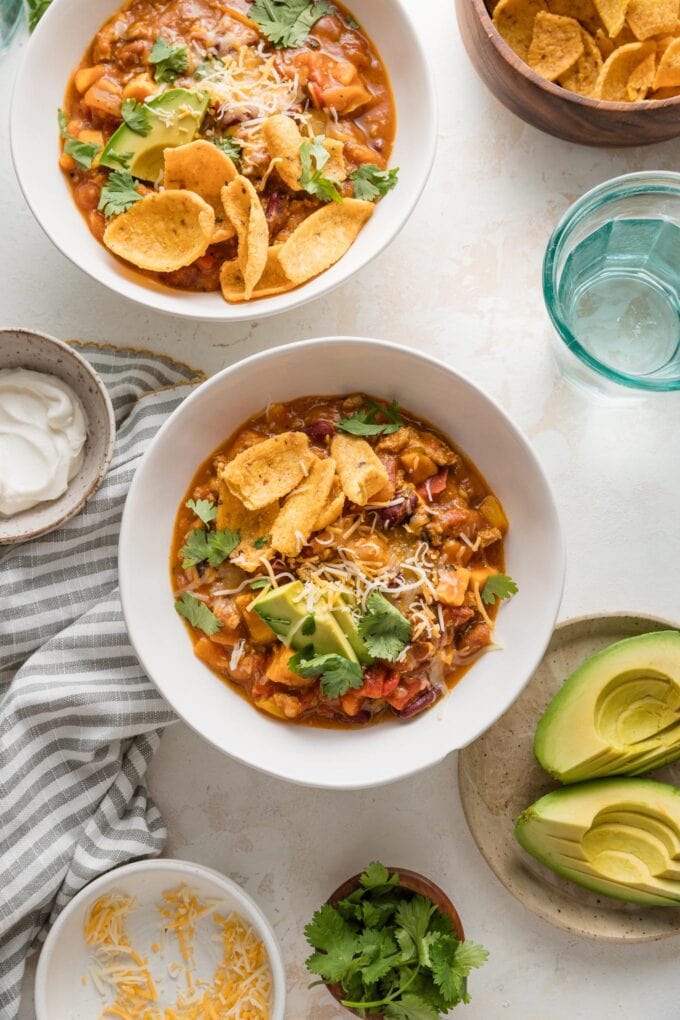 Countertop set with two bowls of pumpkin chili with ground turkey and beans, topped with avocado, corn chips, and sour cream.