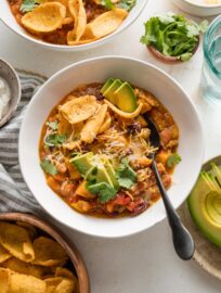 Bowl full of turkey pumpkin chili topped with fresh avocado, corn chips, cheese, and cilantro leaves.