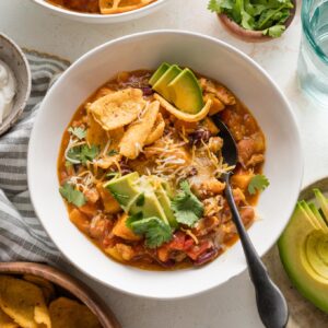 Bowl full of turkey pumpkin chili topped with fresh avocado, corn chips, cheese, and cilantro leaves.