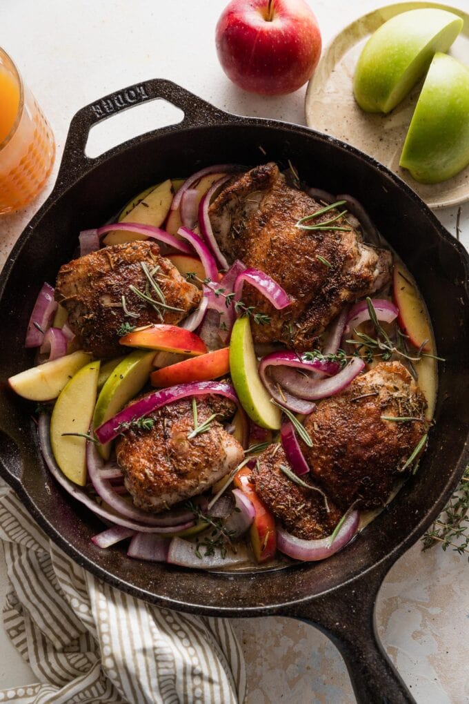 Overhead view of a cast iron skillet filled with pan-seared chicken thighs in an apple cider pan sauce with fresh apples and red onion.
