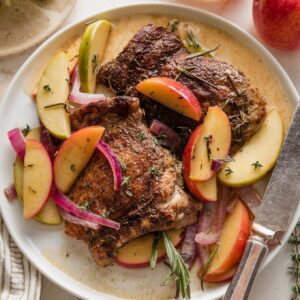 Dinner plate with apple cider braised chicken thigh, sauteed sliced apples and onions, and an apple cider pan sauce topped with fresh rosemary and thyme.