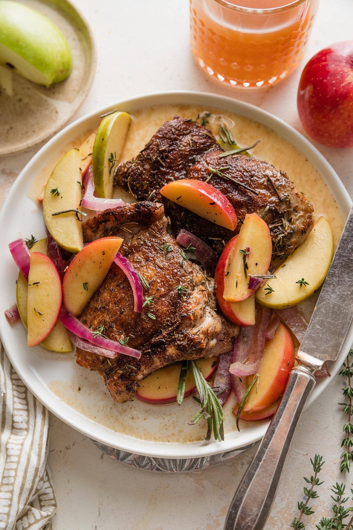 Dinner plate with apple cider braised chicken thigh, sauteed sliced apples and onions, and an apple cider pan sauce topped with fresh rosemary and thyme.