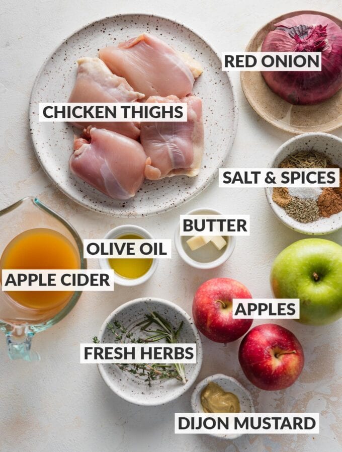 Labeled overhead photo of boneless, skinless chicken thighs, red onion, several apples, fresh apple cider, olive oil and butter, Dijon mustard, salt, spices, and herbs, all measured out and ready to cook.