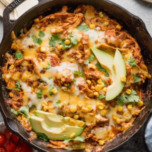 Chicken enchilada skillet dish with corn, green chilies, and red sauce, topped with melted cheese, sliced avocado, and fresh cilantro.