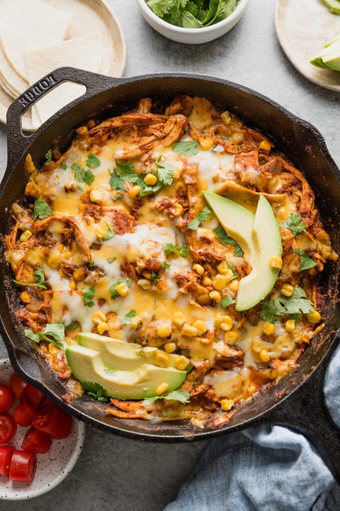 Chicken enchilada skillet dish with corn, green chilies, and red sauce, topped with melted cheese, sliced avocado, and fresh cilantro.