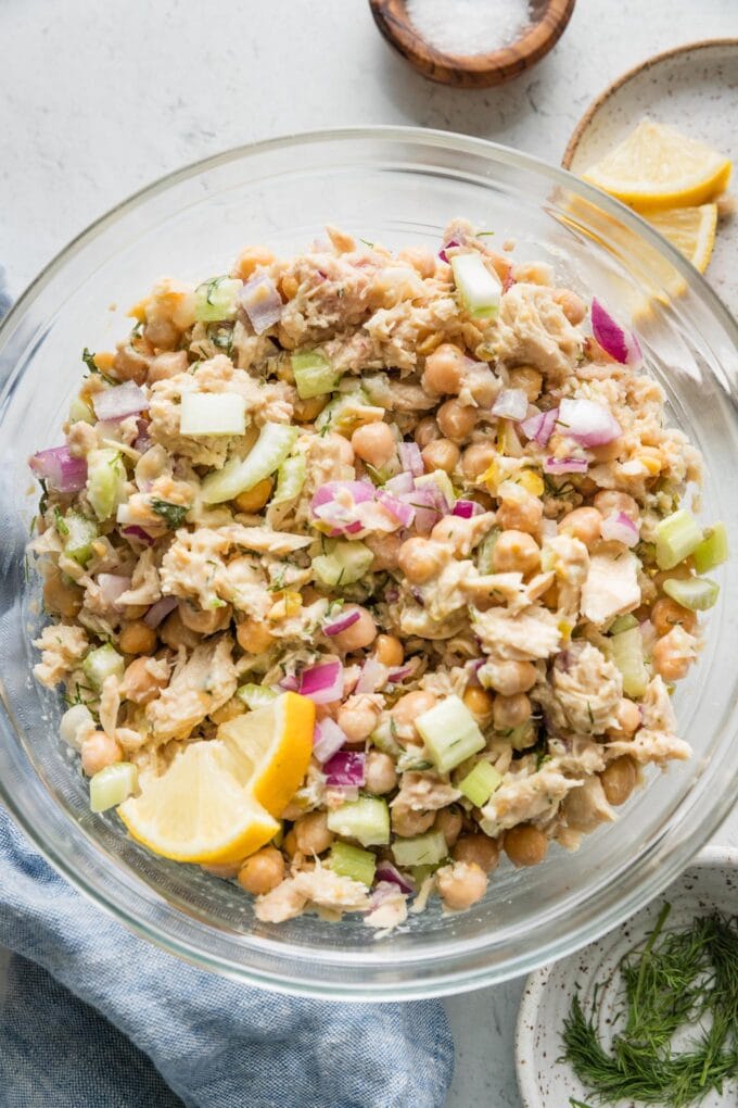 Large glass prep bowl full of chickpea and tuna salad with fresh dill and lemon wedges.