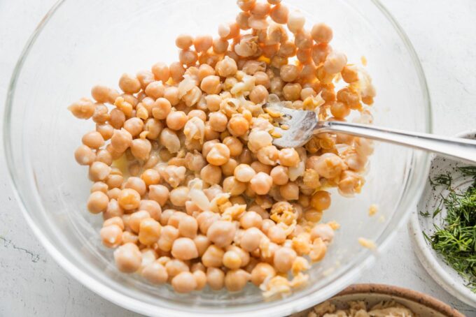 Action shot of some chickpeas being partially mashed with the back of a fork against the side of a large glass bowl.