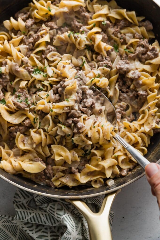 Hand scooping out a generous serving of easy ground beef and noodles from a large skillet.