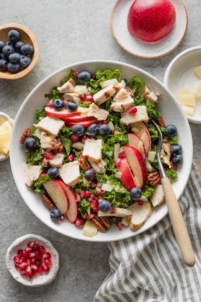Countertop set with a large salad of kale, apples, chicken, and berries, with extra pomegranate seeds, cheese, and blueberries in prep bowls nearby.