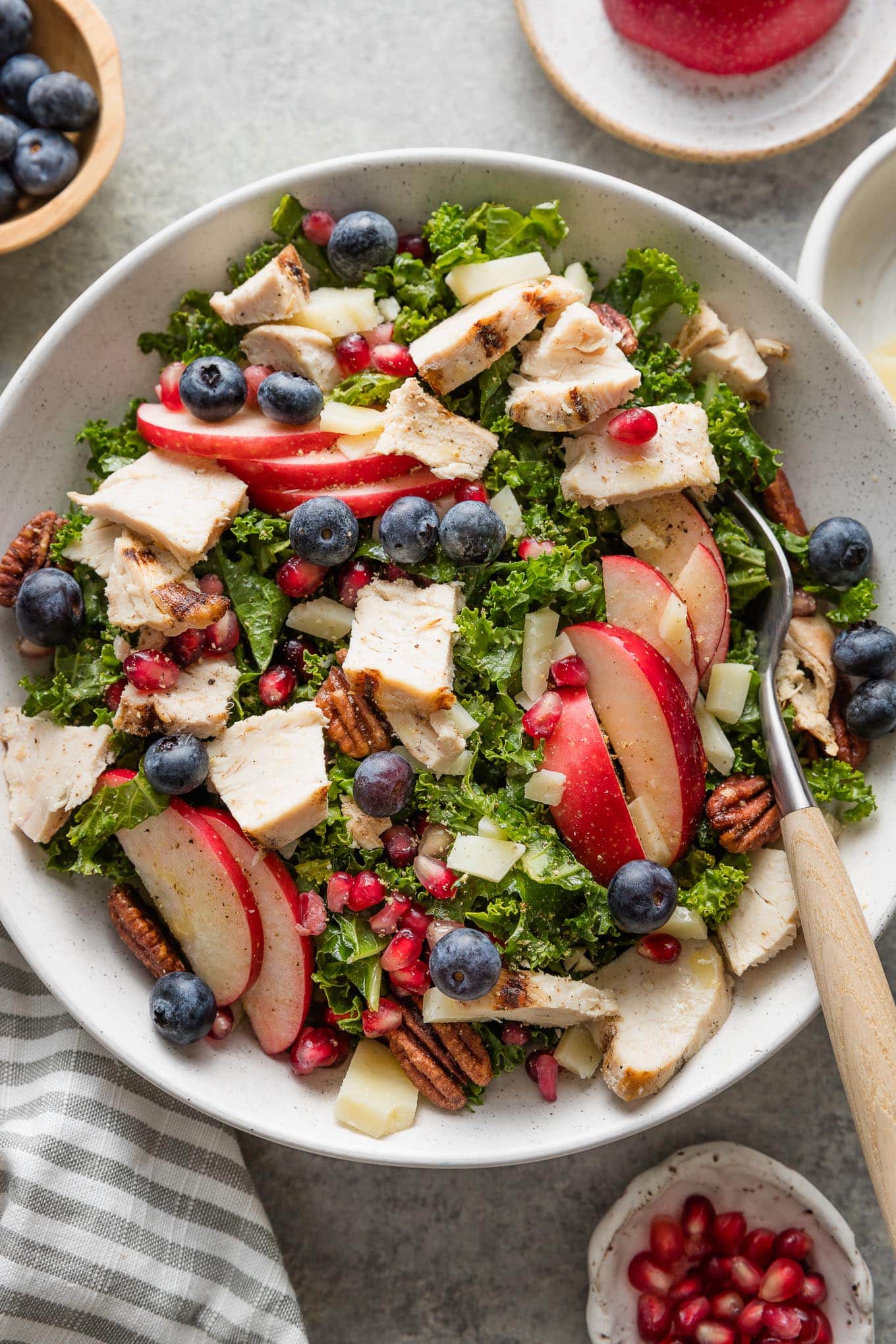 Bowl full of a meal-sized kale salad with chopped apples, fresh blueberries and pomegranate arils, Pecorino, pecans, and grilled chicken.