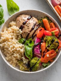 Bowl with a balsamic-marinated chicken thigh, roasted veggies, and pearl couscous, all drizzled with balsamic glaze and topped with fresh basil leaves.
