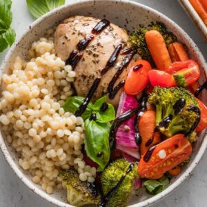 Bowl with a balsamic-marinated chicken thigh, roasted veggies, and pearl couscous, all drizzled with balsamic glaze and topped with fresh basil leaves.