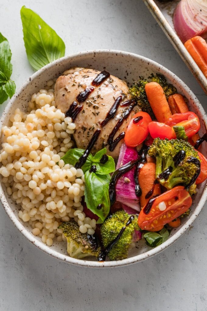 Bowl with a balsamic-marinated chicken thigh, roasted veggies, and pearl couscous, all drizzled with balsamic glaze and topped with fresh basil leaves.