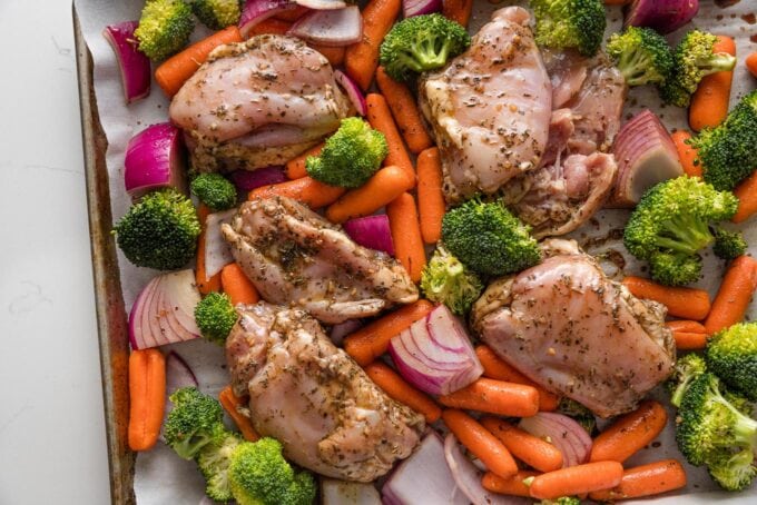 Sheet pan with balsamic marinated chicken thighs, broccoli florets, red onion, and baby carrots, about to go into the oven.