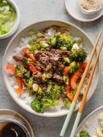 Bowl of sheet pan beef and broccoli served over rice with garnishes of green onions and sesame seeds on top, with chopsticks positioned to the side of the bowl.