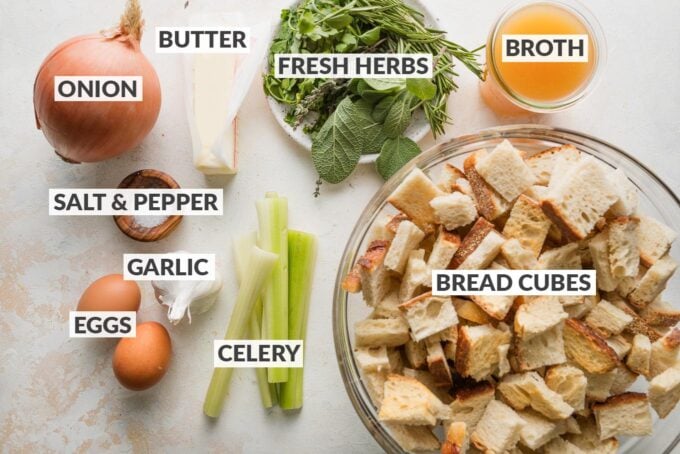 Labeled overhead photo of bread cubes, celery, onion, fresh herbs, chicken broth, eggs, salt, and pepper.