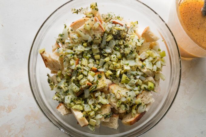 Onion-herb mixture poured over bread cubes in a very large glass prep bowl.