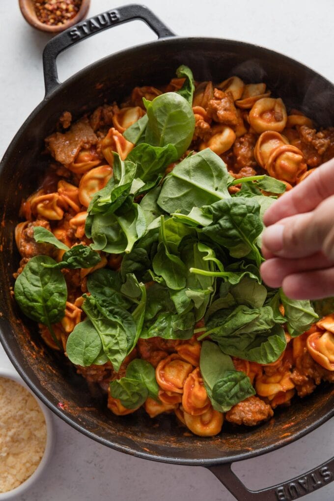 Action shot of hand adding baby spinach to wilt into a dish of tortellini with tomato sauce and sausage.