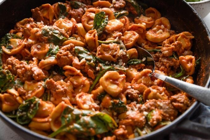 Large cast iron skillet full of tortellini with sausage in tomato cream sauce, with a serving spoon dipped in and about to lift out a helping.