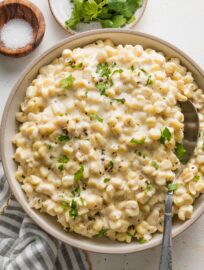 Bowl of creamed corn garnished with fresh parsley, with extra greens and a pinch bowl of salt nearby on the countertop.