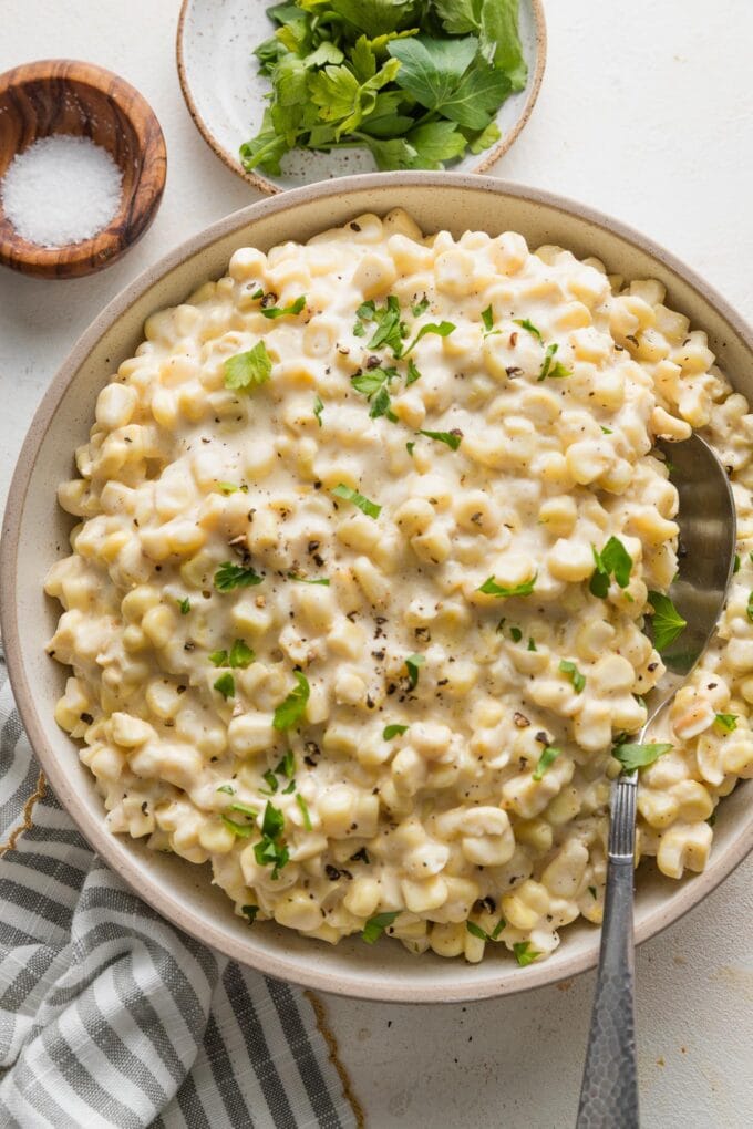 Bowl of creamed corn garnished with fresh parsley, with extra greens and a pinch bowl of salt nearby on the countertop.