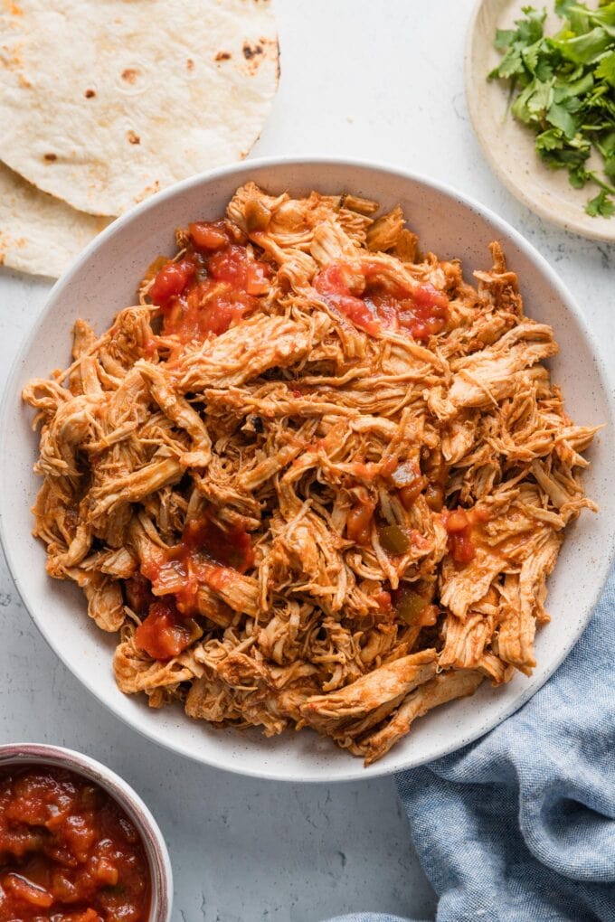 Overhead view of a serving bowl full of Instant Pot salsa chicken with extra salsa, chopped fresh cilantro, and tortillas nearby on the counter.