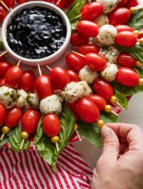 Close up of a woman's hand picking up a Caprese skewer from a platter arranged to look like a wreath.