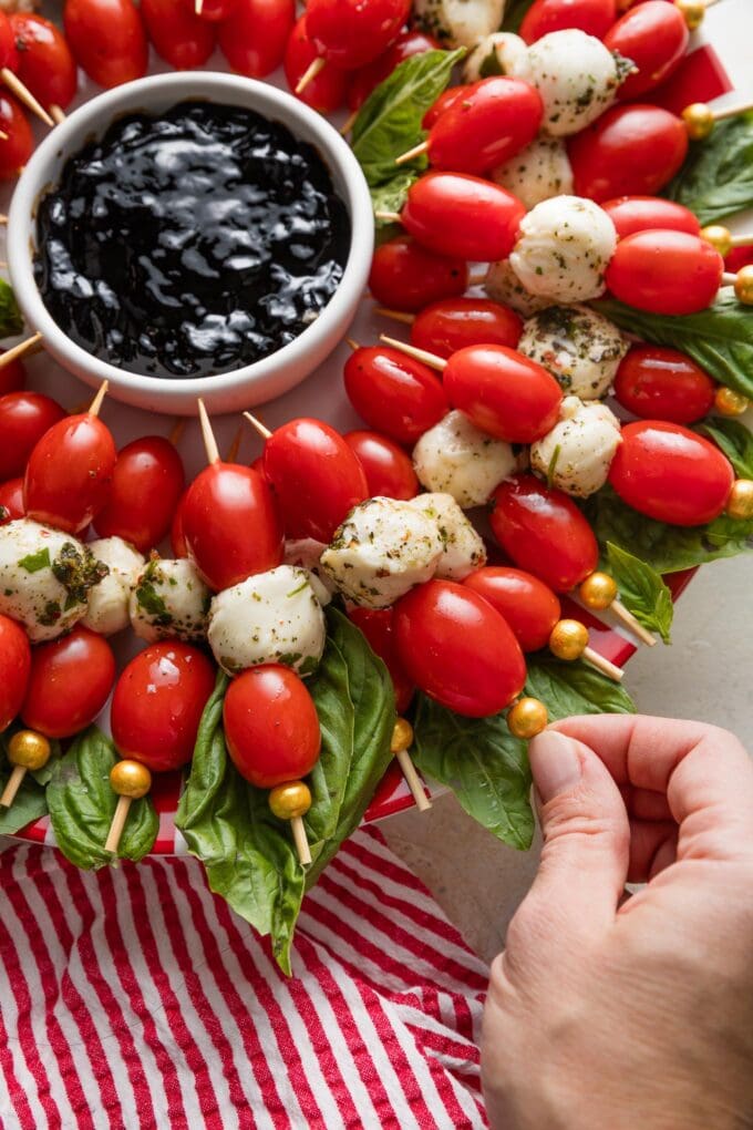 Close up of a woman's hand picking up a Caprese skewer from a platter arranged to look like a wreath.