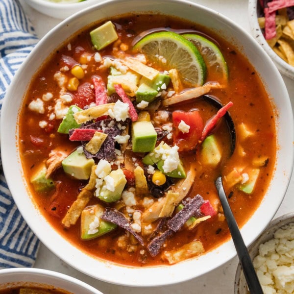 Overhead view of a bowl of chicken chipotle tortilla soup topped with tortilla strips, avocado, and lime wedges.