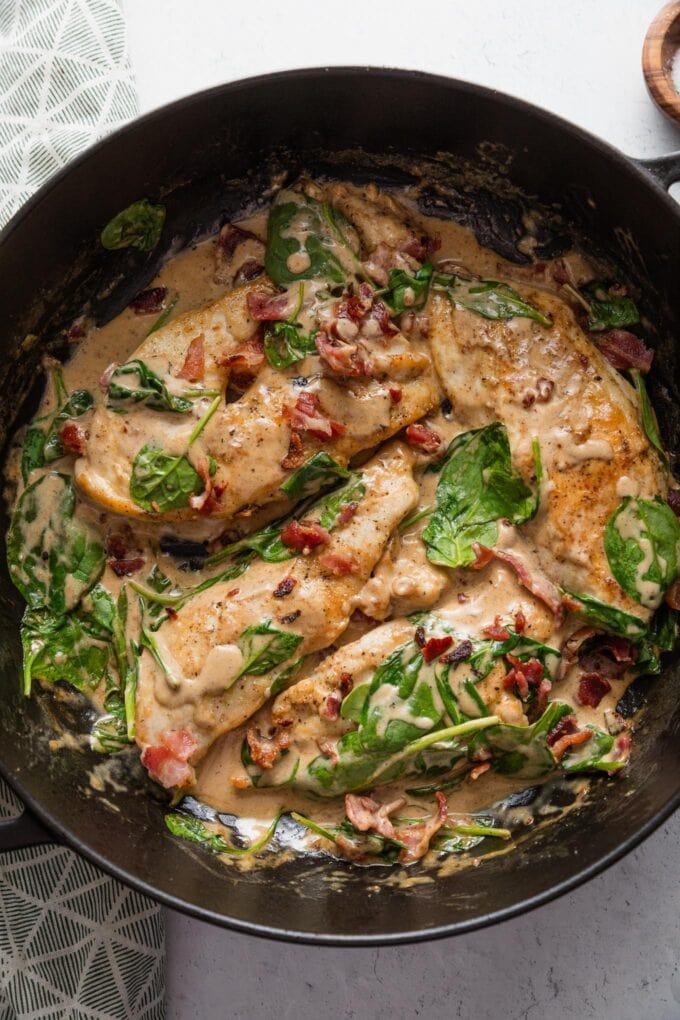 Overhead view of a deep cast iron skillet with chicken breasts, bacon, baby spinach, and a light balsamic cream sauce.