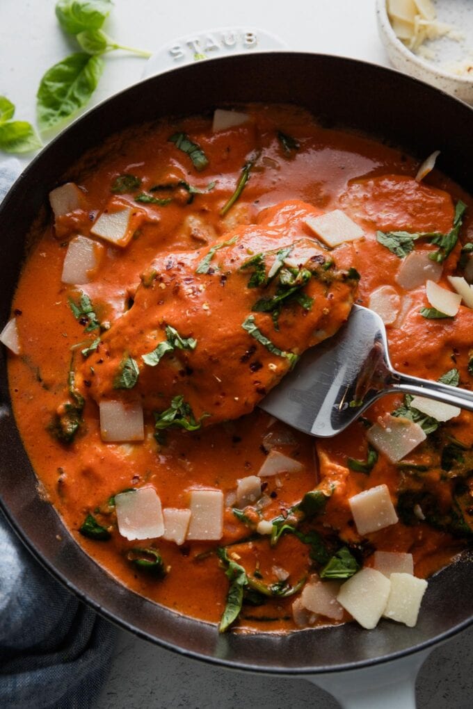 Silver serving utensil lifting up a chicken breast from a pan of chicken in homemade tomato cream sauce.