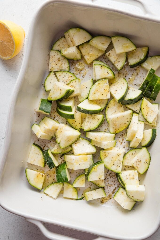 Chopped zucchini in a white baking pan sprinkled with salt, pepper, and olive oil.