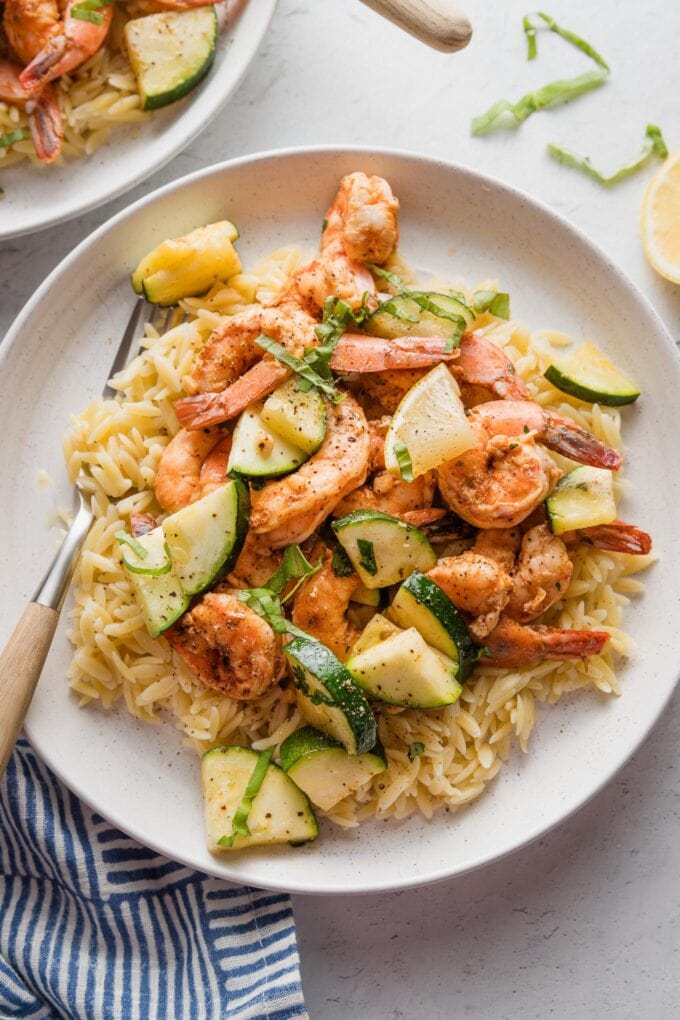 Plate of orzo, shrimp, and zucchini, all served with fresh herbs and lemon wedges.