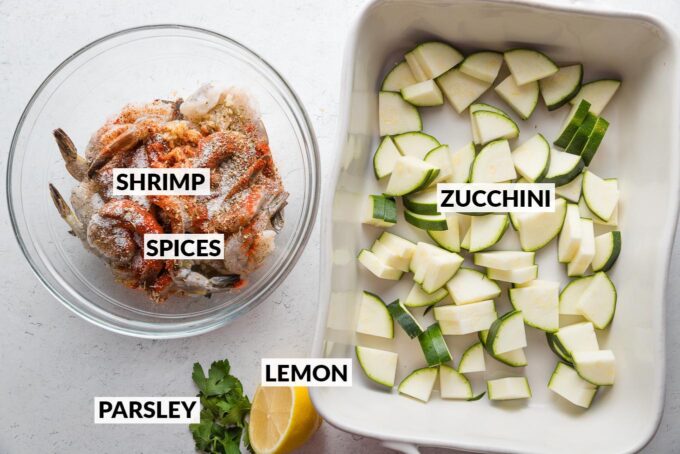 Labeled ingredient photo of chopped zucchini, uncooked shrimp seasoned with a blend of olive oil, lemon juice, and spices, and half a fresh lemon and parsley leaves.