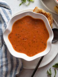 Scalloped white bowl full of a super quick and creamy homemade tomato soup with grilled cheese wedges and fresh slivered basil scattered on the counter nearby.