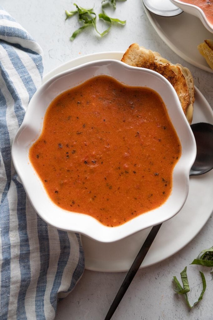 Scalloped white bowl full of a super quick and creamy homemade tomato soup with grilled cheese wedges and fresh slivered basil scattered on the counter nearby.