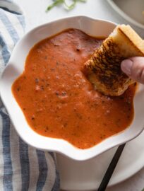 Close up of a woman's hand dunking a grilled cheese sandwich into a bowl of homemade tomato soup in a scalloped white bowl.
