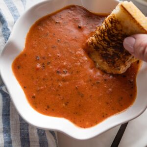 Close up of a woman's hand dunking a grilled cheese sandwich into a bowl of homemade tomato soup in a scalloped white bowl.