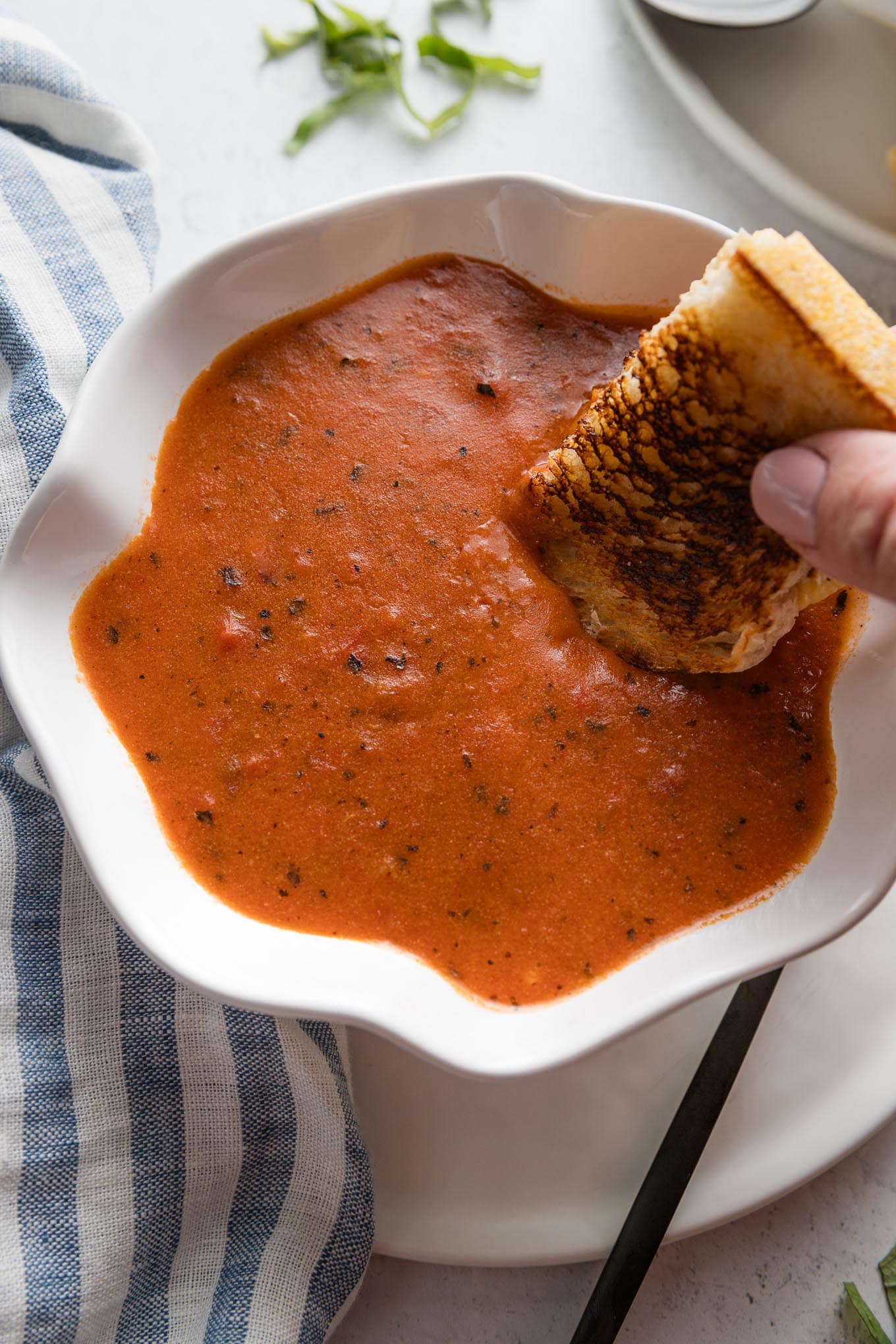 Close up of a woman's hand dunking a grilled cheese sandwich into a bowl of homemade tomato soup in a scalloped white bowl.