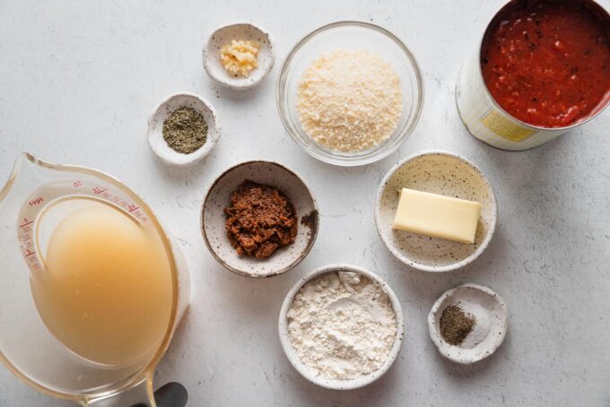 Landscape flat lay photo of ingredients for an ultra-quick homemade tomato soup: high quality canned crushed tomatoes, low sodium chicken broth, sun-dried tomato pesto for extra flavor, dried basil, salt, and pepper, Parmesan cheese, garlic, butter, and all-purpose flour for a quick roux.