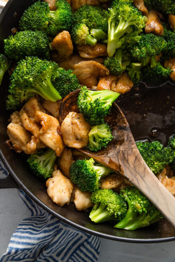 Close up of a wooden spoon lifting a portion of chicken and broccoli stir fry out of a pan.