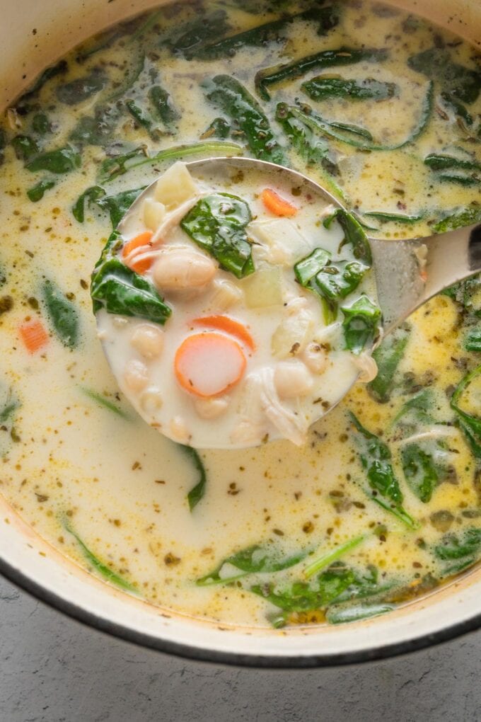Close up view of a ladle scooping up a serving of chicken soup with pasta and white beans from a Dutch oven.