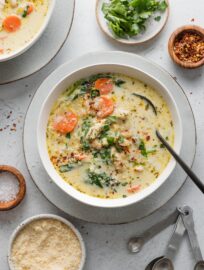 Counter set with bowls of a light yet creamy chicken soup with carrots and pasta, with extra parsley, red pepper flakes, and Parmesan in bowls nearby to scatter on top.