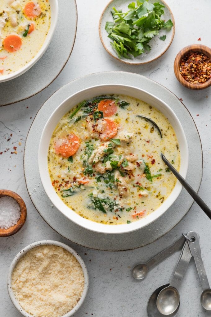 Counter set with bowls of a light yet creamy chicken soup with carrots and pasta, with extra parsley, red pepper flakes, and Parmesan in bowls nearby to scatter on top.