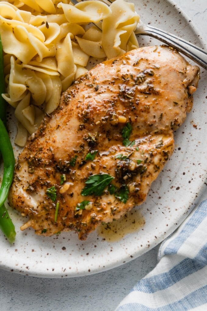 Close up of a garlic butter chicken breast with herbs served with egg noodles and green beans on a speckled ceramic dinner plate.