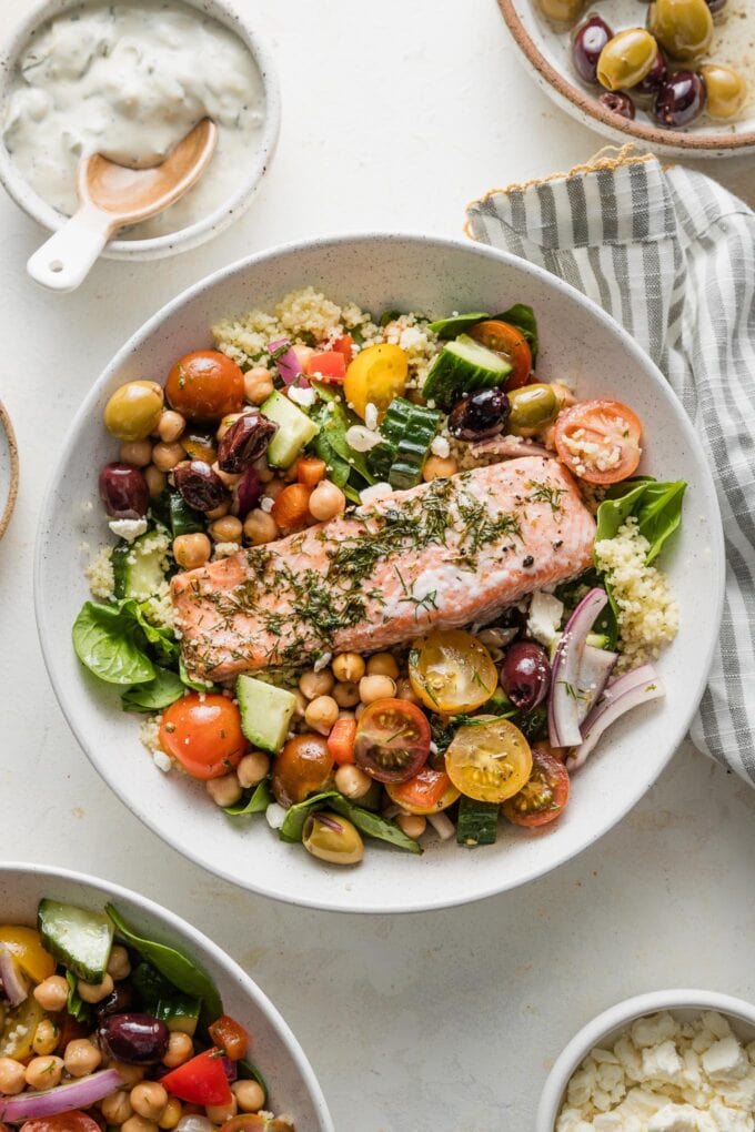 Countertop set with a big Greek salmon grain bowl, plus extra herbs, couscous, and Tzatziki sauce for garnish.
