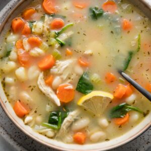 Close up of a bowl of white bean soup with veggies, mini gnocchi, and shredded chicken, with fresh parsley and a lemon wedge sprinkled on top.